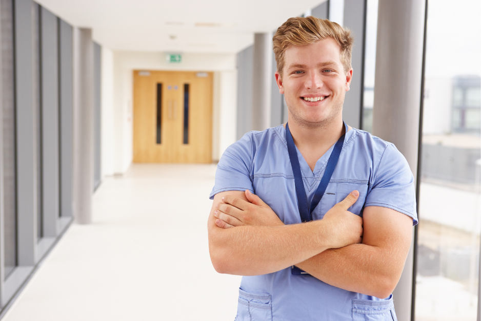 Eine lächelnde männliche Pflegeperson steht mit verschränkten Armen im Gang eines Krankenhauses.
