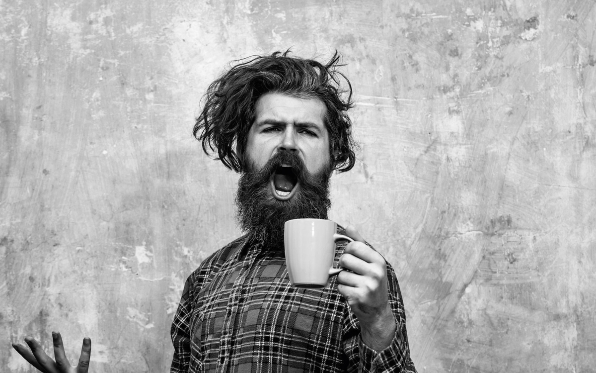 Ein Mann mit lockigem, zerzaustem Haar und Bart, hält eine Tasse in der linken Hand und singt.