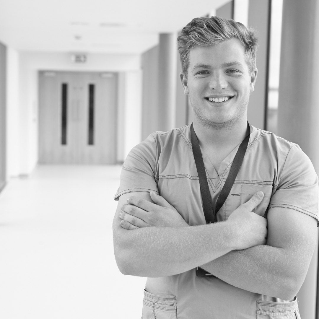 Eine lächelnde männliche Pflegeperson steht mit verschränkten Armen im Gang eines Krankenhauses.