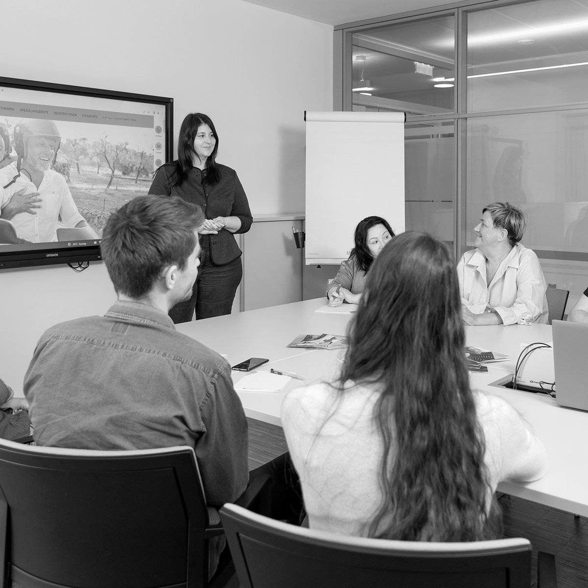 Sechs Personen sitzen um einen Konferenztisch und eine Person steht vorne vor einem Flipchart und einem Bildschirm mit einer Präsentation.